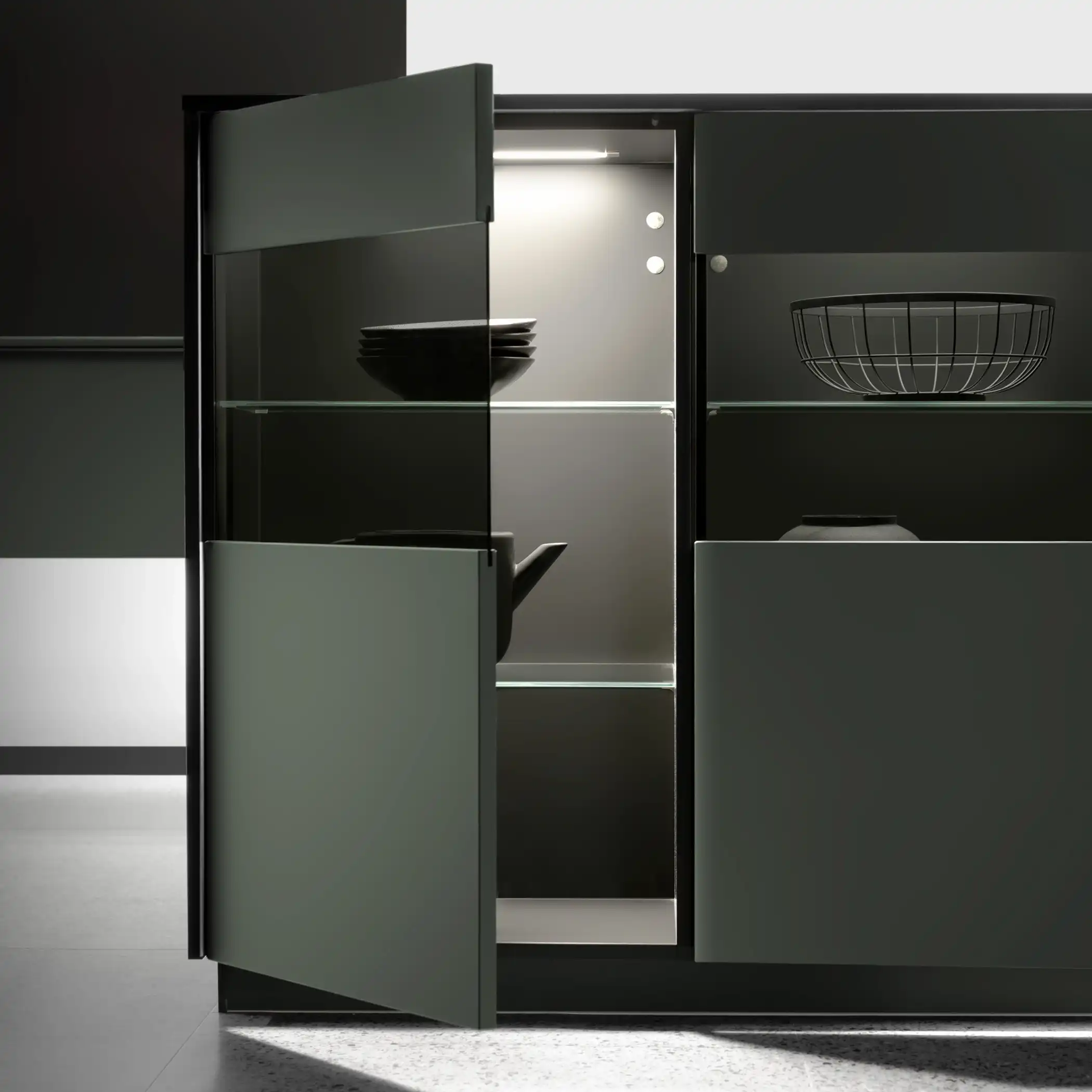 Easytouch Mineral Green and BlackGlass A close-up view of a modern, high-end kitchen cabinet in a deep green finish, partially open to reveal neatly arranged shelves. The cabinet features an illuminated interior showcasing stacked plates, various kitchen tools, and a basket. The sleek design and subtle lighting highlight the cabinet’s sophisticated look and functionality.